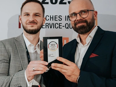 Široka fotografija prikazuje pozornicu za dodjelu nagrada iza publike koja sjedi. Na lijevoj strani pozornice, muškarac u tamnom odijelu i žena u dugoj bijeloj haljini stoje s tabletima u rukama, okrenuti prema publici. U središtu tamne pozadine pozornice, veliki ekran prikazuje svjetleći jantarni tekst na vrhu na kojem piše "WIZJA I INNOVACJE". Ispod toga, centrirano na ekranu, nalazi se velika, svjetleća kristalna nagrada u obliku dijamanta, nakon čega slijedi tekst "Maciej Góralski" i "Vasco Electronics" ispod. Topla, difuzna svjetla osvjetljavaju pozornicu nasuprot mračnom, prepunom auditoriju.