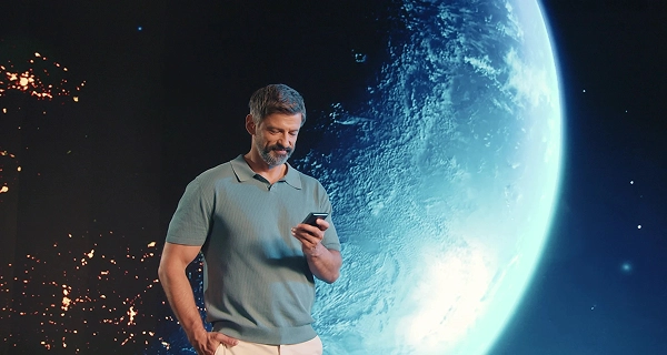 A photograph of a middle-aged man with a salt-and-pepper beard and hair, wearing a light blue short-sleeved polo and white trousers. He stands with his right hand in his pocket, looking down at the translator held in his left hand. The background is a massive, curved screen displaying a deep space panorama: a large, cold-blue Earth dominates the right side, while glowing golden nebulae and distant city-like lights dot the dark void on the left, along with scattered stars. The blue light from the Earth projection casts a hue on his side.
