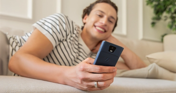 A close-up photograph focusing on a person's hand holding a dark blue handheld device with a VA logo on the back. In the softly blurred background, a smiling young man wearing a black and white striped t-shirt lies relaxed on a light-colored couch. The setting is a bright, modern living room with soft lighting and a hint of indoor greenery in the background.