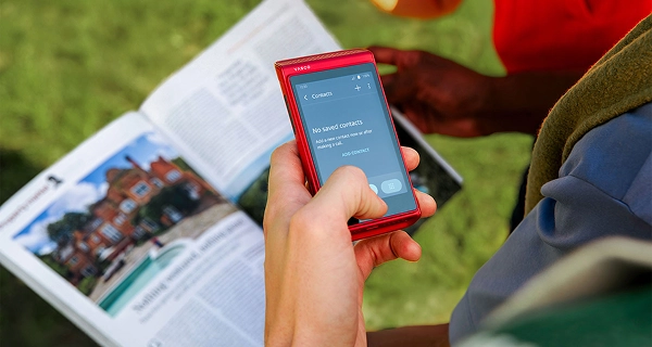 A close-up photograph of a hand holding a red Vasco electronic device over an open travel guidebook or brochure. The device's screen displays a "Contacts" menu showing the text "No saved contacts. Add a new contact now or after making a call." alongside an "ADD CONTACT" option. In the background, a blurry green lawn is visible, along with the arm of another person wearing a red shirt.