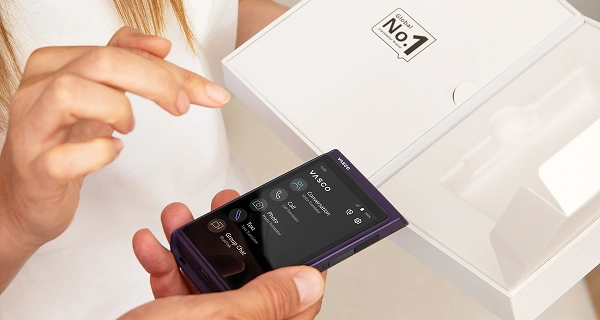 A close-up photograph of two people interacting with a new device. One person holds a dark purple handheld electronic device that displays a numeric dialing keypad on its screen. A woman with long brown hair wearing a white top stands next to them, reaching out to touch or point at the screen. In the background, an open white product box is held up, displaying a graphic logo that reads "No. 1" on the inside of the lid.
