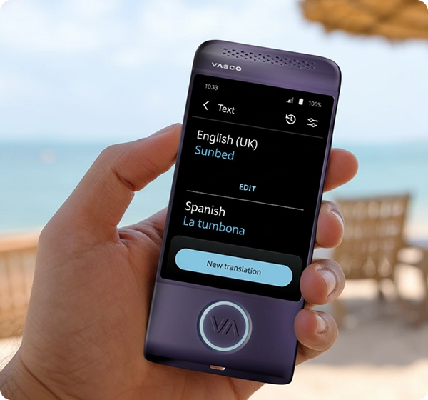 Krupni plan ruke koja drži ljubičasti elektronički prevoditelj Vasco. Na zaslonu se prikazuje tekstualni prijevod s engleskog (UK) na španjolski, a riječ "Sunbed" (ležaljka za sunčanje) prevedena je kao "La tumbona" ​​(ležaljka za sunčanje). Pozadina je svijetla, sunčana plaža s meko fokusiranim pogledom na ocean, zlatni pijesak, nekoliko ležaljki i slamnati suncobran. 