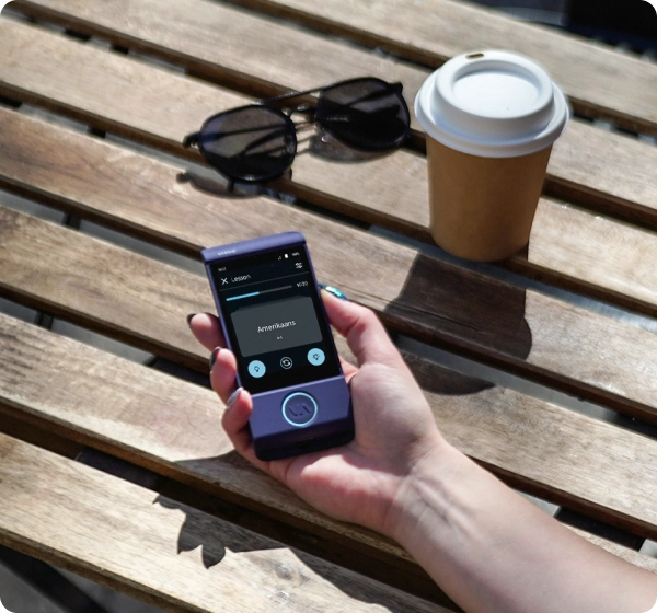 A person's hand holding a small, purple handheld translator device over a wooden slatted table. The screen shows a language lesson with the word 